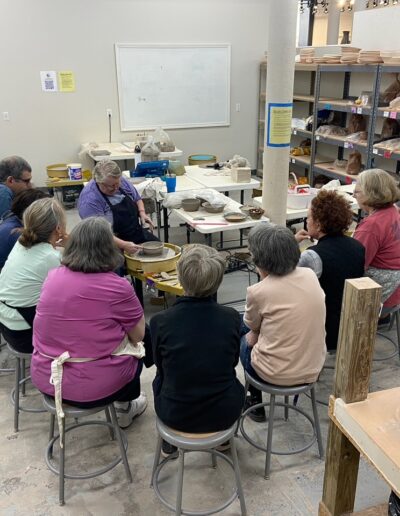 Julia Mann demonstrating at potters wheel, surrounded by students, teaching center, intermediate wheel class