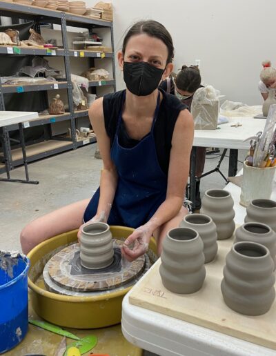 Teaching Center student happy while throwing vase on the potters wheel, sitting next to her other leather hard pots, intermediate wheel class