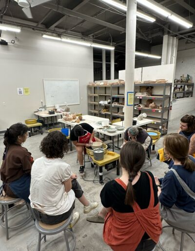 Katie Meili Messersmith demonstrating throwing a bowl on the potters wheel, surrounded by students, intermediate wheel class 2