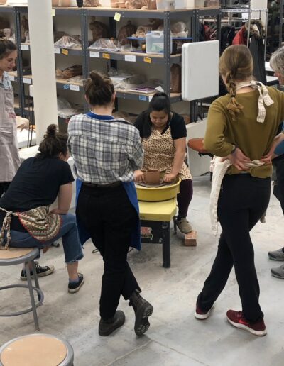 Katie Meili Messersmith demonstrating at the potters wheel for her intermediate wheel class, students watching